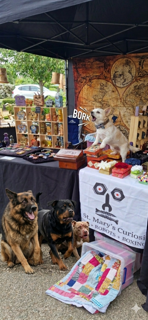 A lively scene at a craft fair stall for Jon and Mary's Curios, featuring a small dog wearing a bowtie standing on boxes and 'borking,' with two larger dogs sitting faithfully in front of the table. The stall is decorated with an old-world map backdrop and displays several of the 'CUTIES'—the handmade wooden robot figures—on shelves, while the table is covered in a banner featuring a mechanical skull logo